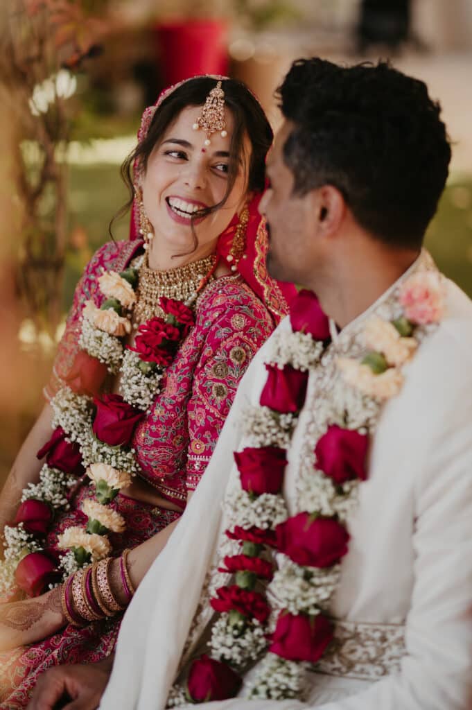 Mariée indienne souriante en lehenga rose et lourds bijoux, portant une varmala de roses avec son marié en tenue blanche.