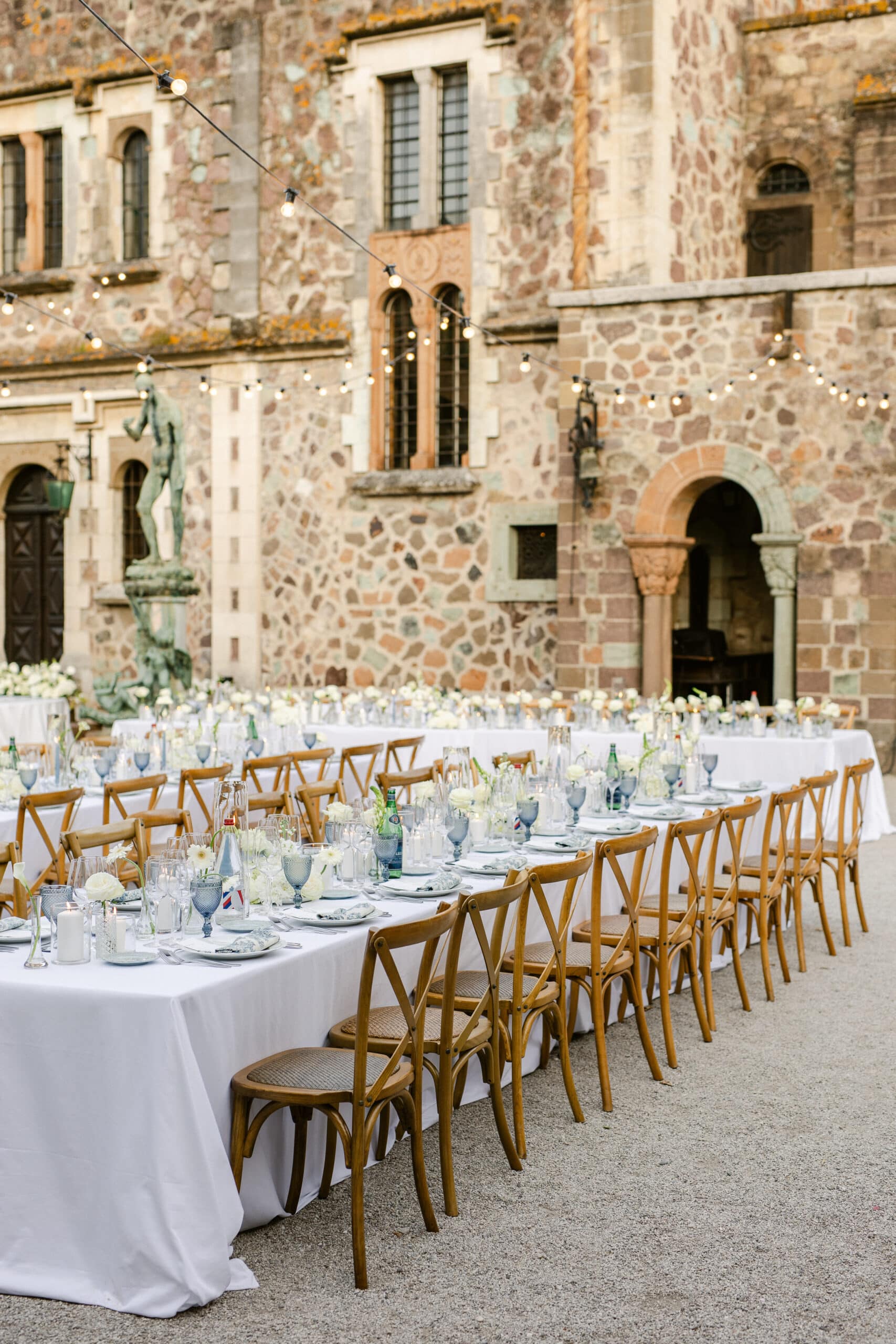 Longue table de réception de mariage extérieure élégante devant un château en pierre. Chaises bistrot, décor fleuri blanc et guirlandes lumineuses.