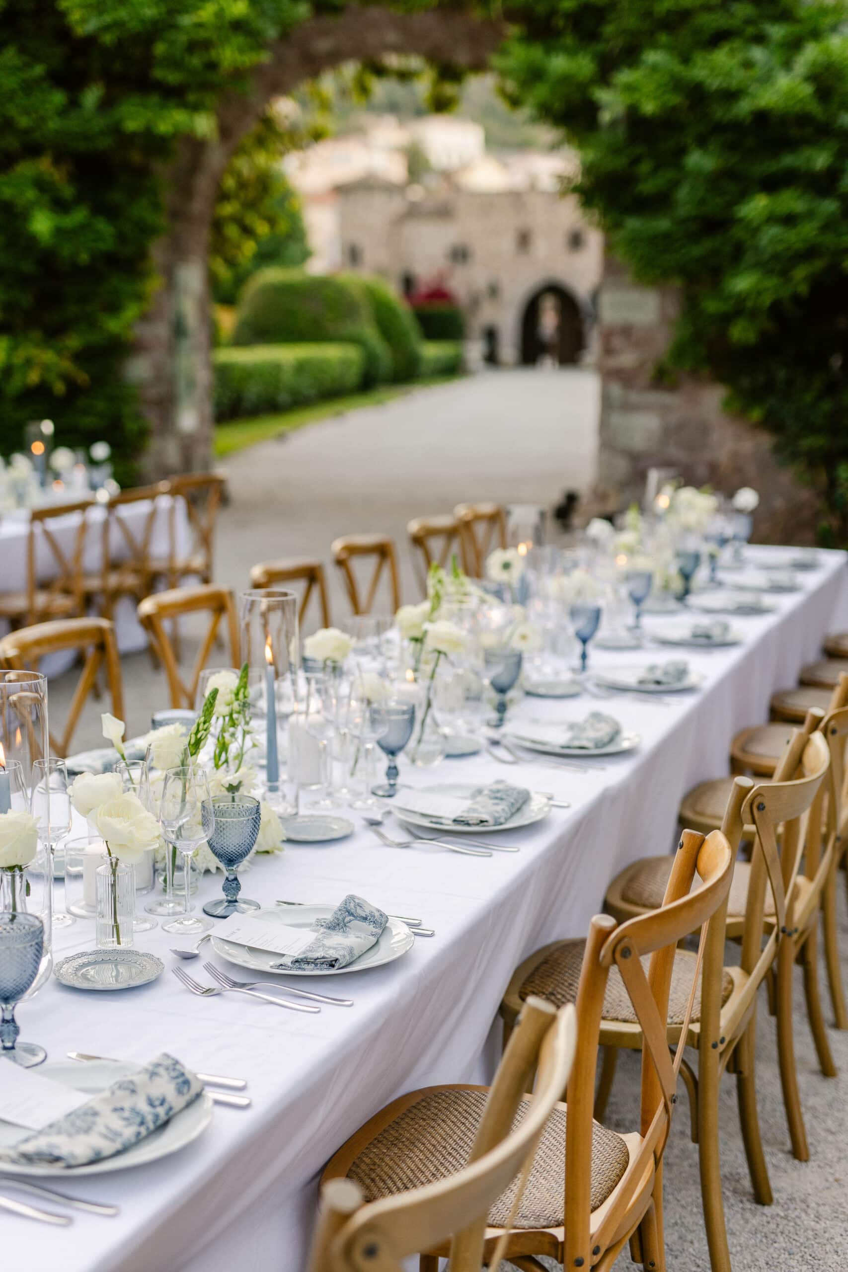 Dîner de mariage en plein air. Longue table de réception avec nappe blanche, chaises bois, centres de table de roses crème et vaisselle bleu-gris dans un jardin de château.