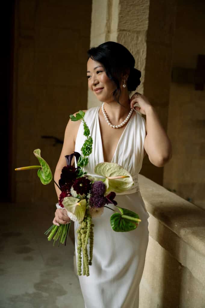 Portrait d'une mariée élégante en robe blanche, collier de perles, souriant. Elle tient un bouquet moderne d'Anthuriums verts sur fond de pierre.