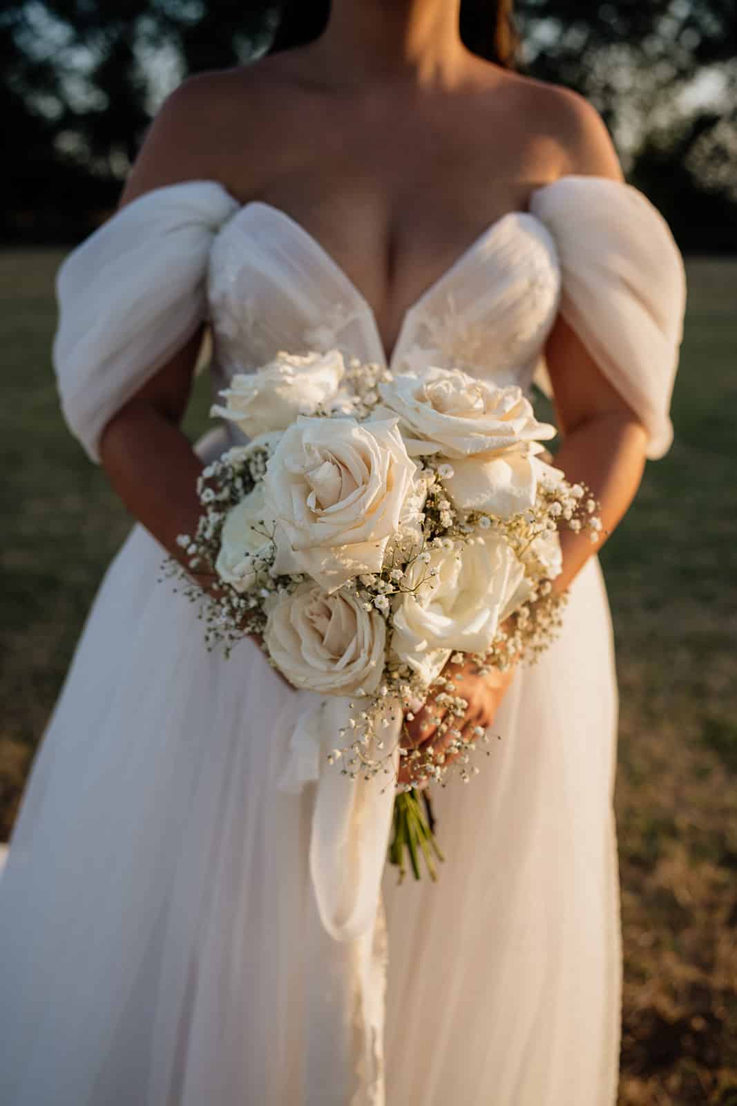 Mariée en robe blanche à épaules dénudées tenant un bouquet dense de roses crème et de gypsophile sous la chaude lumière dorée.