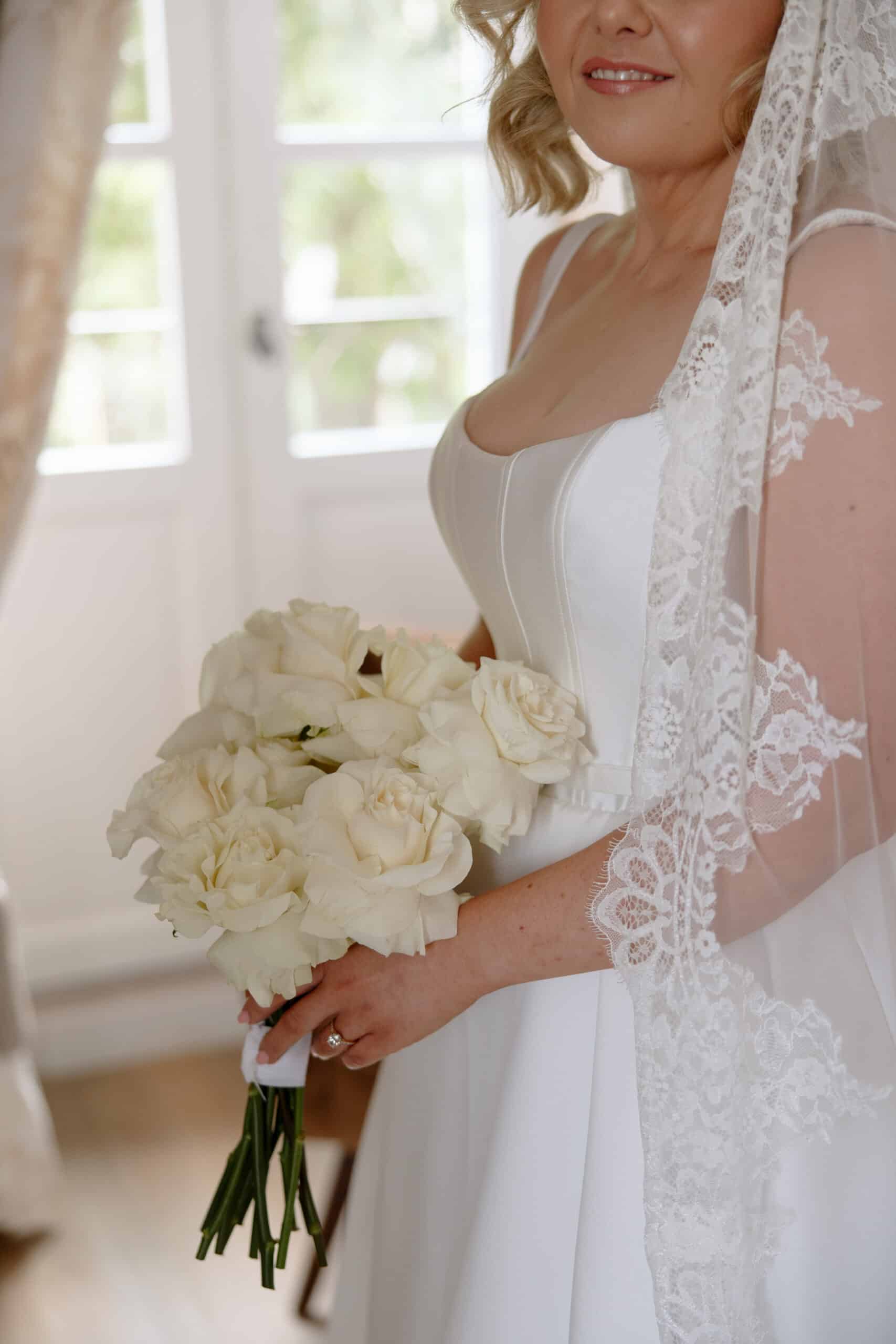 Mariée en robe de satin blanc, souriant légèrement, tenant un bouquet de roses blanches. Voile en dentelle élégant.