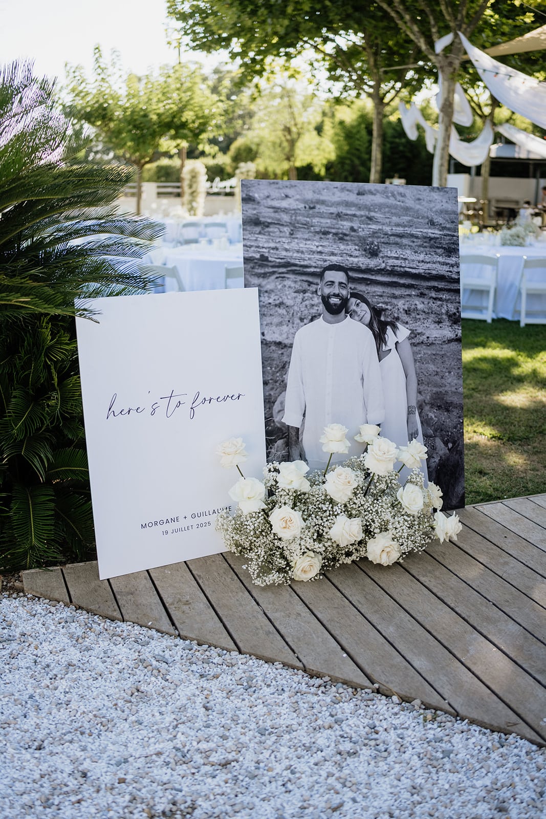 Panneaux de bienvenue de mariage : portrait B&W Morgane et Guillaume, "here's to forever" (19 juillet 2025). Décor extérieur élégant, roses blanches et gypsophile.