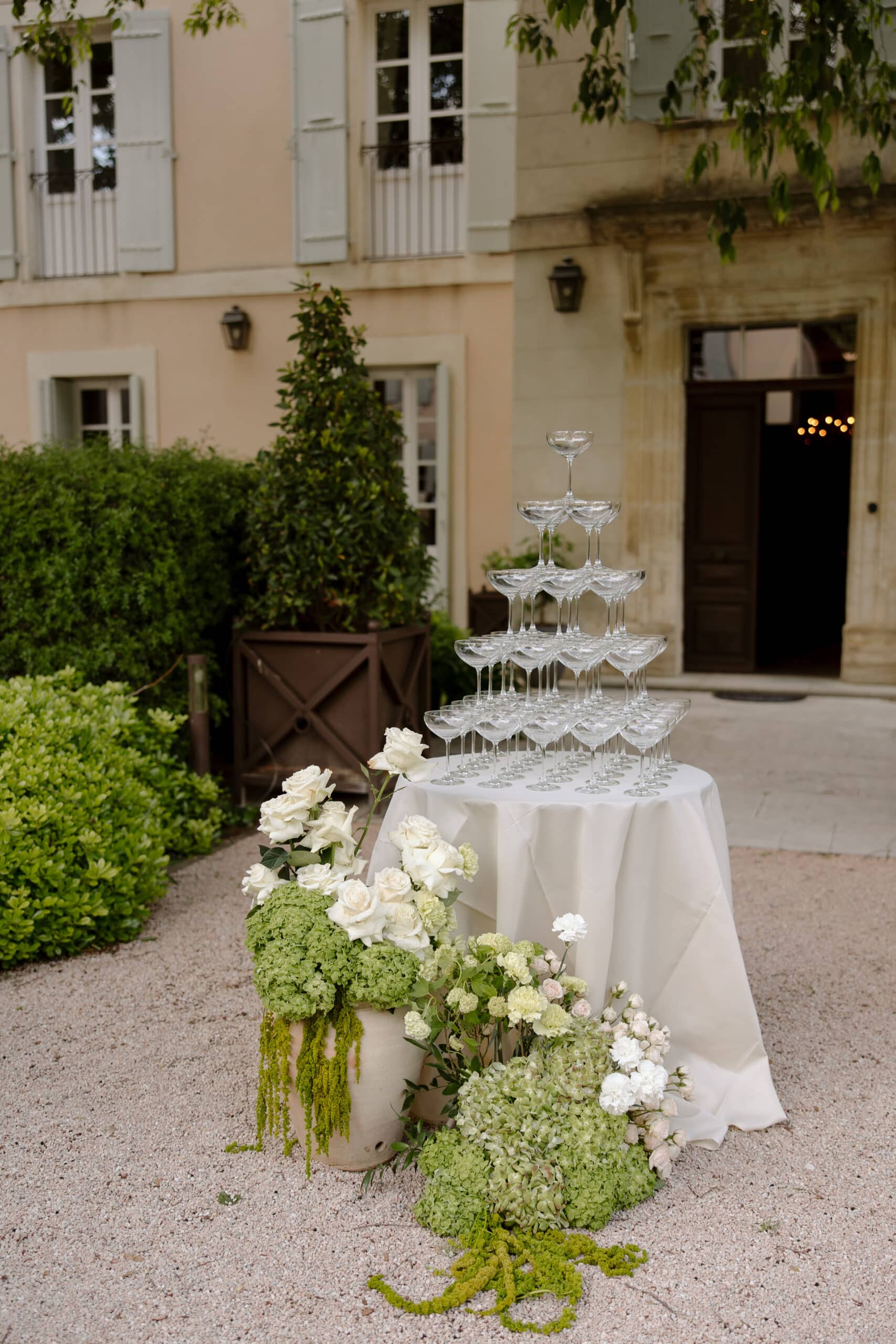Réception de mariage dans un jardin : pyramide de coupes de champagne et arrangements floraux blancs et verts devant une villa.