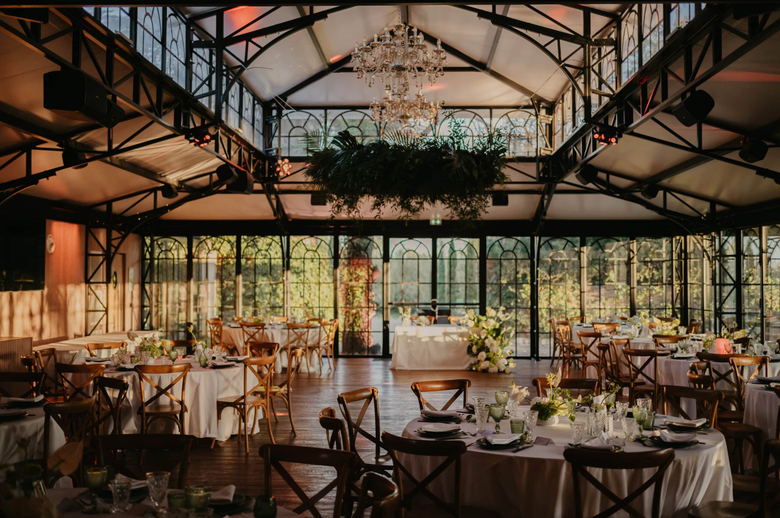 Serre de réception élégante pour mariage. Tables dressées, chaises bois, chandeliers, et grand décor végétal suspendu sous les poutres métalliques.