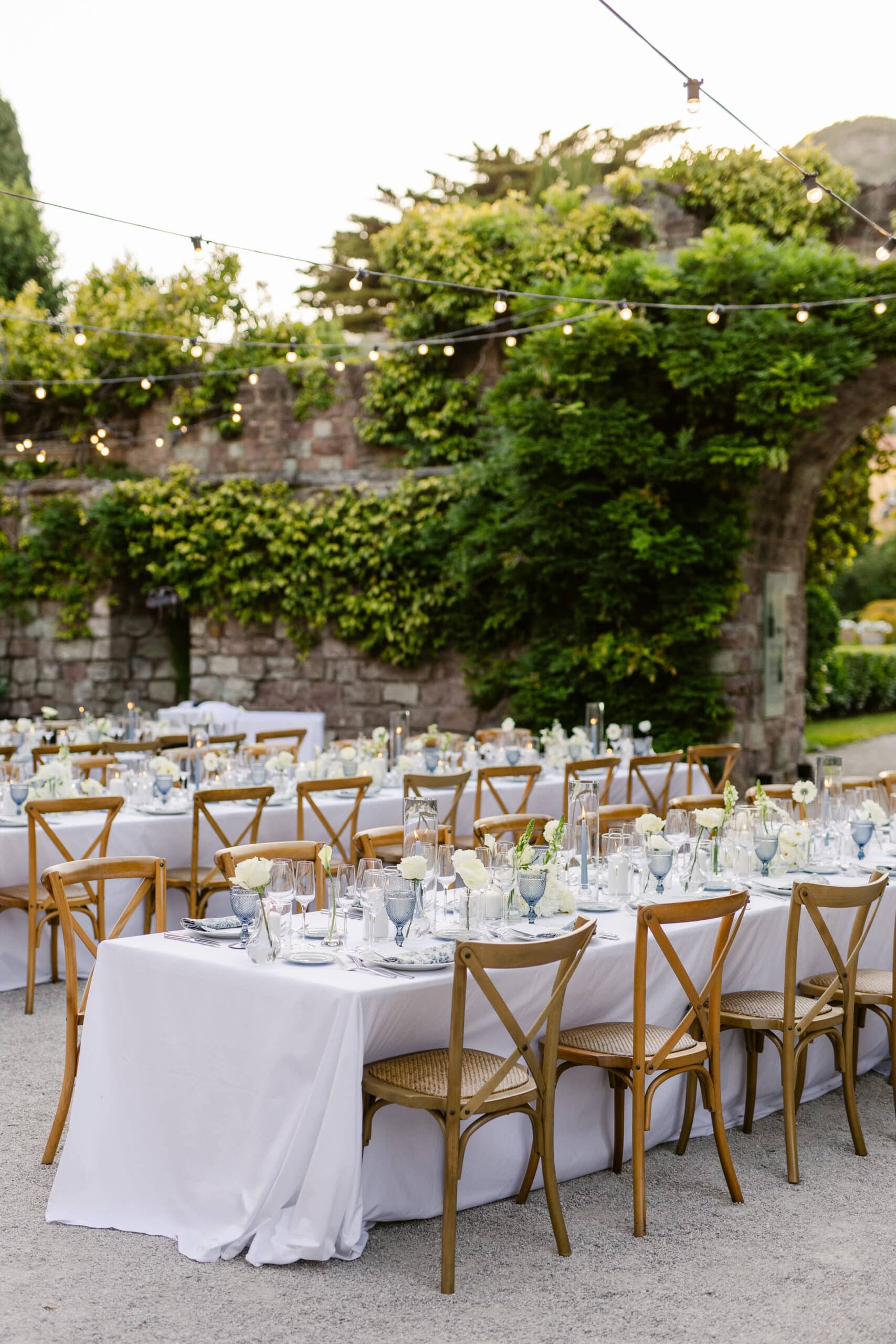 Longues tables de mariage au crépuscule. Murs de pierre couverts de lierre, guirlandes lumineuses, chaises en bois, fleurs et verrerie bleue.