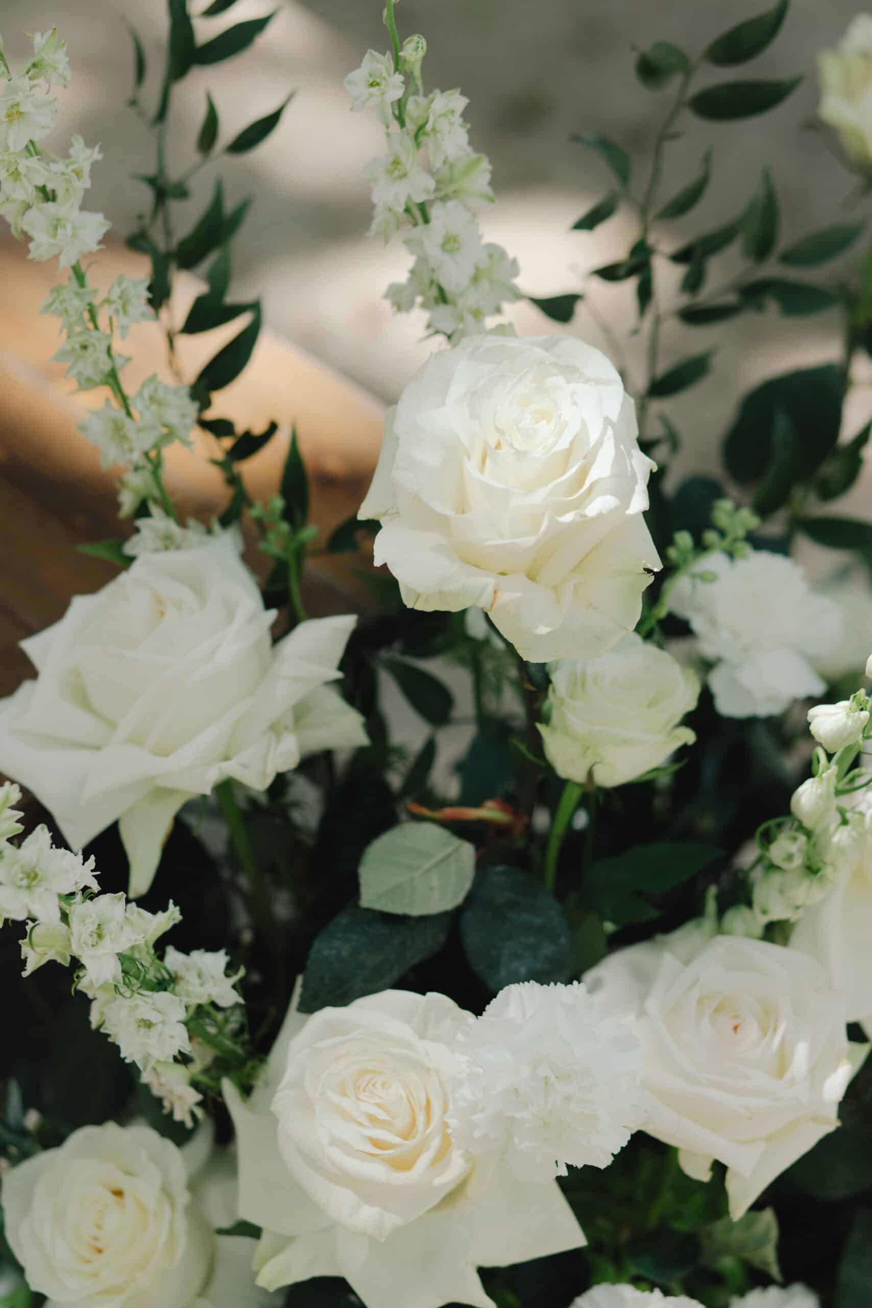 Gros plan sur un arrangement de mariage de roses blanches, de delphiniums et de feuillage vert foncé sous une lumière naturelle.