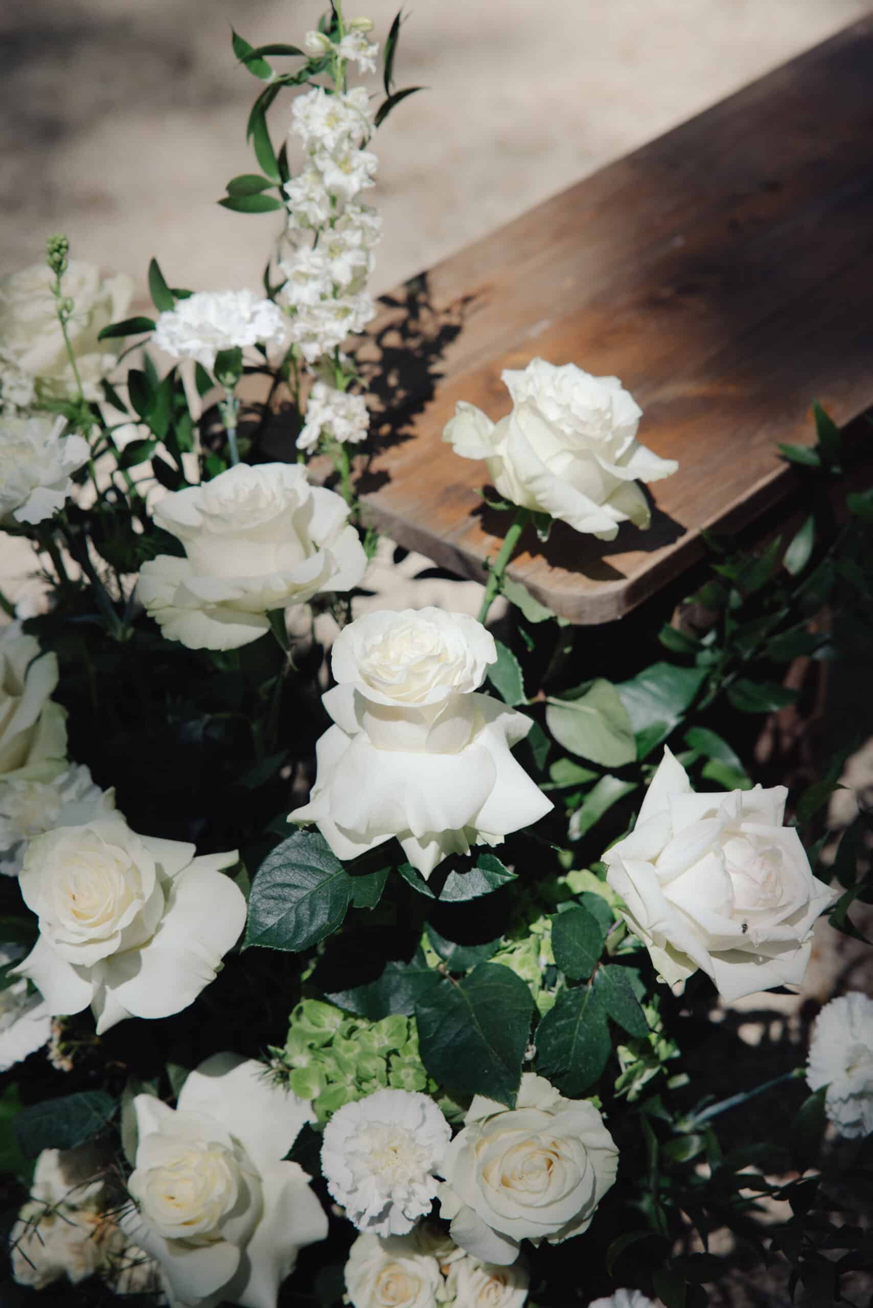 Composition élégante de roses blanches éclatantes et de delphinium, avec feuillage vert luxuriant, près d'un plateau en bois foncé en extérieur.