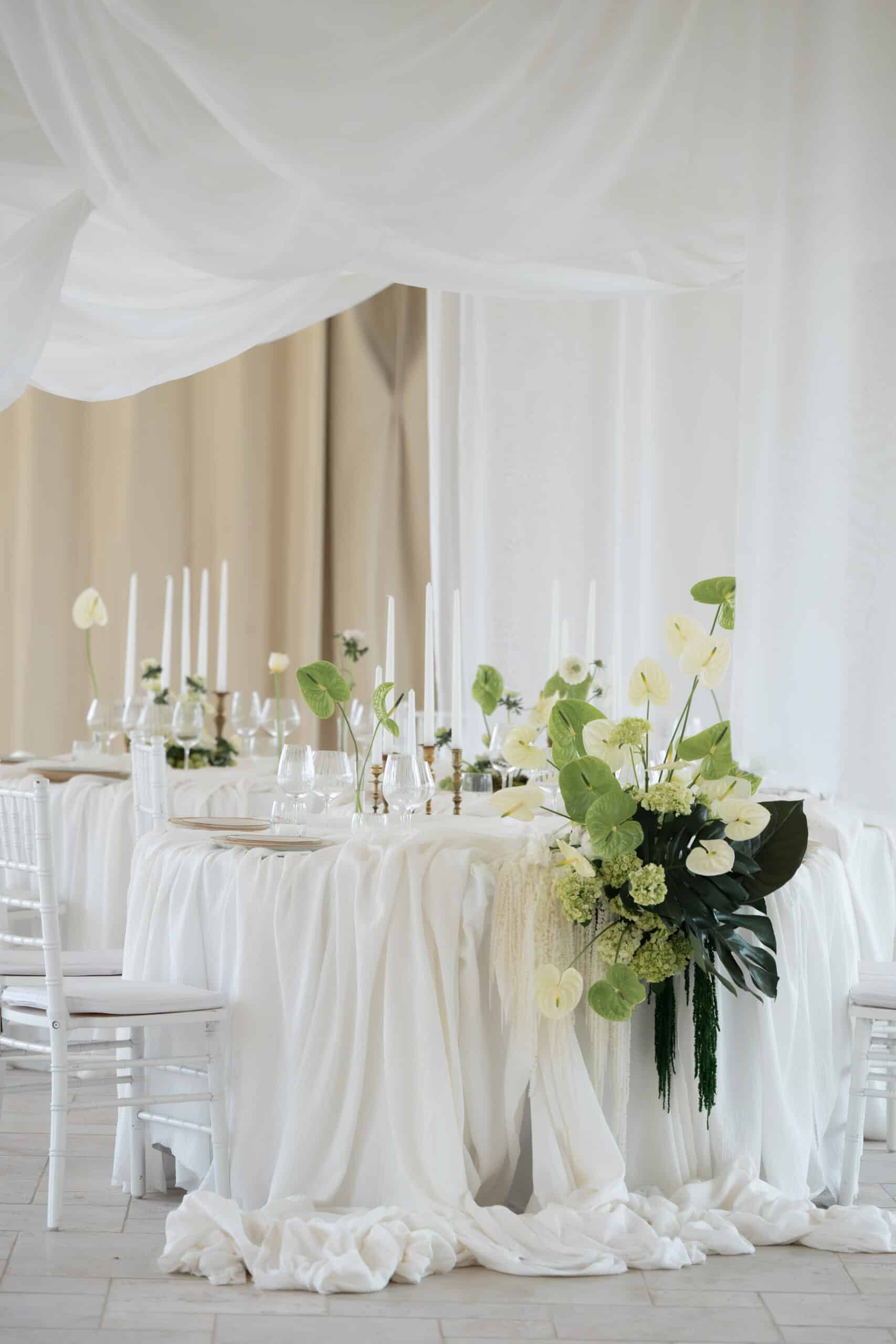 Décoration de table de mariage blanche et verte. Longue table drapée, fleurs modernes (Anthurium) et bougies. Ambiance chic sous voilages.