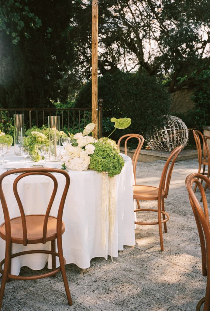 Élégante table de mariage en extérieur : nappe blanche, chaises bistrot en bois, centre de table floral vert et blanc.